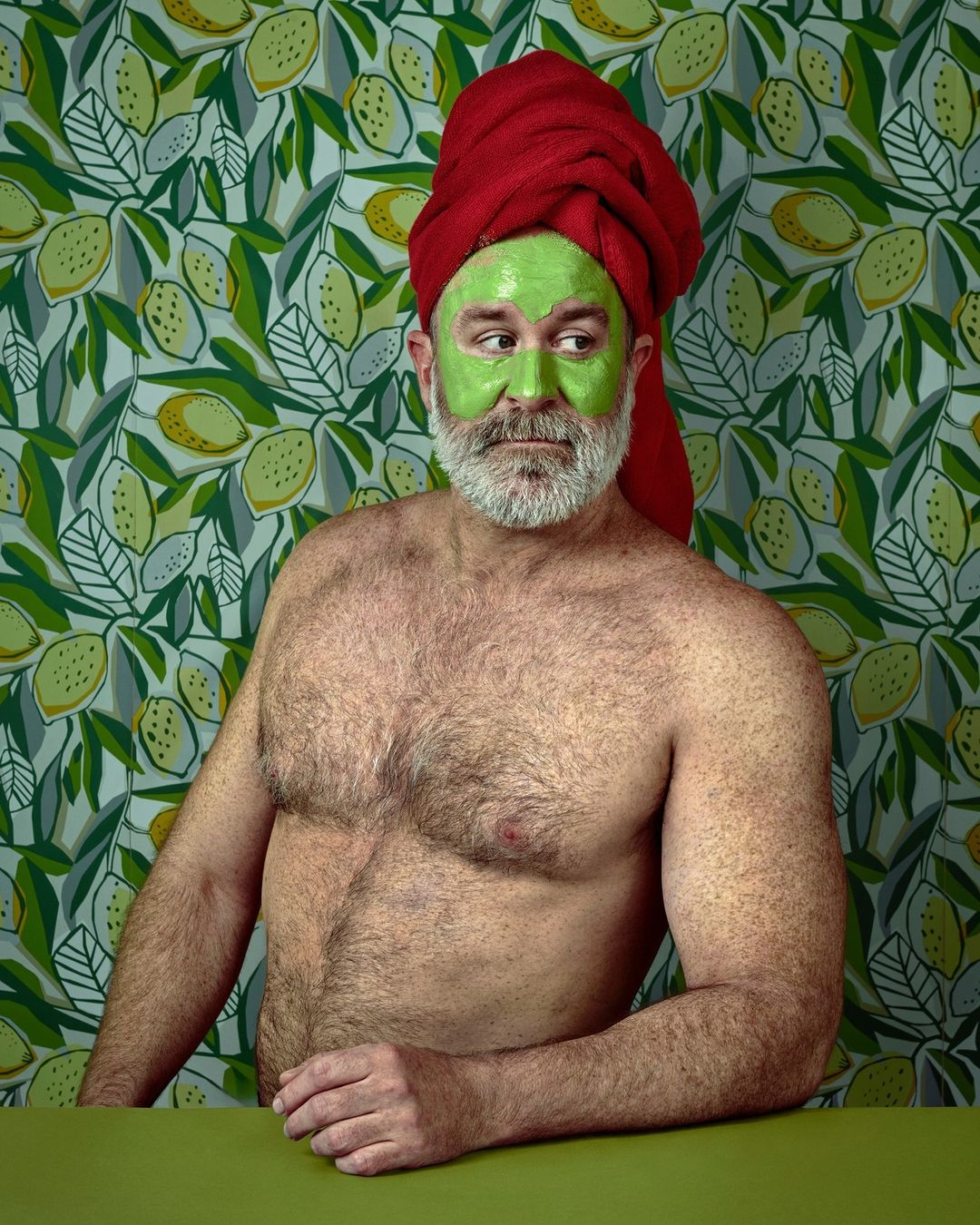 A man with a beard and a red towel on his head, wearing a green facial mask, poses in front of a lemon-patterned wallpaper.